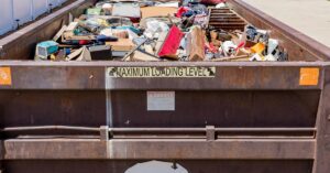 A brown dumpster parked in a driveway, filled to the brim with debris, trash, and other items from a home cleanout or renovation.
