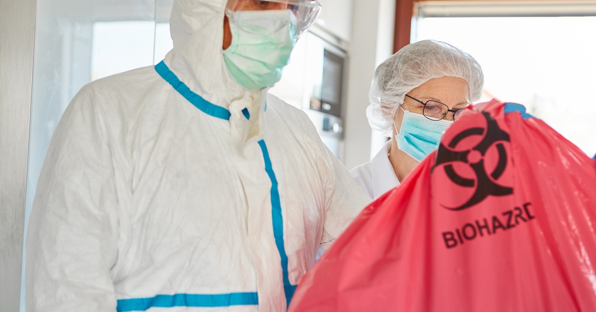 A close-up of a man and woman wearing protective masks and bodysuits while holding a red bag marked Biohazard.