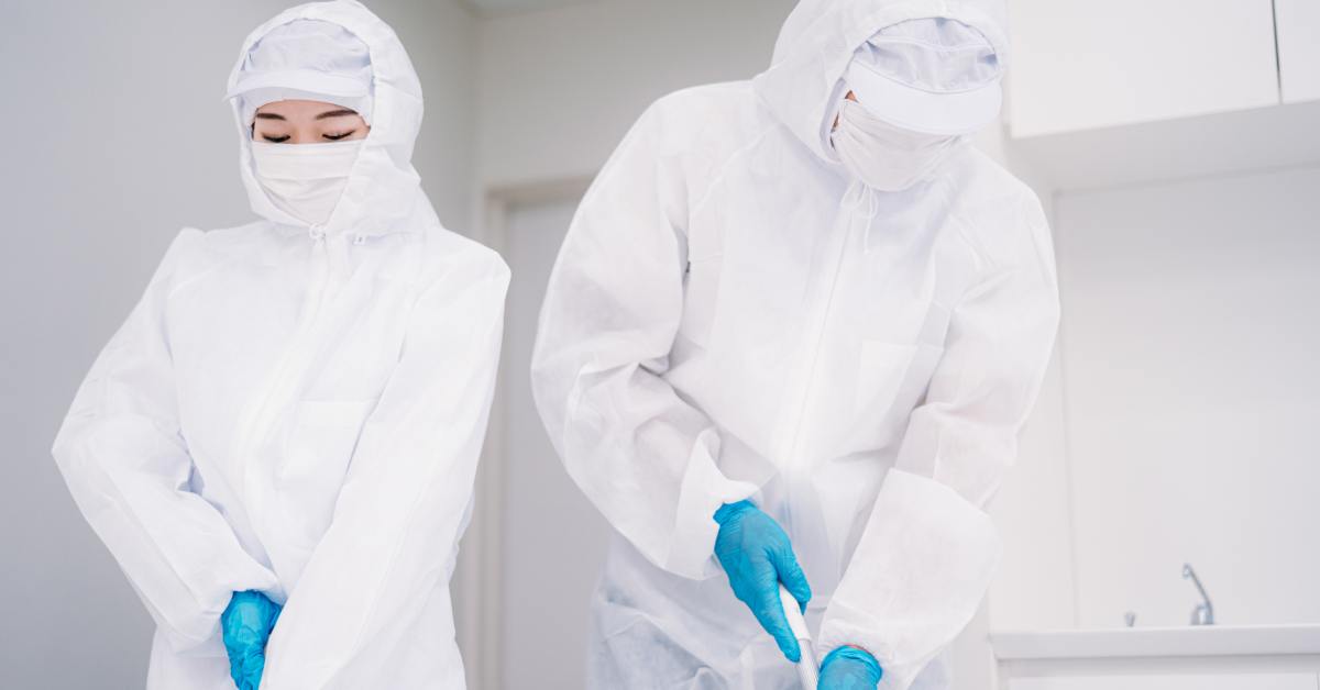 Two people wearing full-body white protective suits and only their eyes showing clean a white room with extending tools.