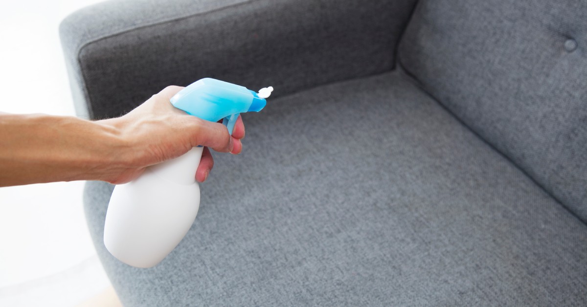 A close-up on a person cleaning a gray couch with a white spray bottle with a blue spray nozzle.