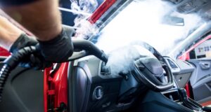 A man's arms wearing gloves hold a stream cleaner, steaming the interior of a red with heavy steam, bilowing inside.