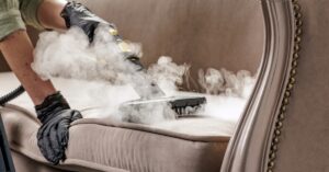 A professional cleaner wearing gloves cleans the beige upholstery on a couch with a steam machine brush.