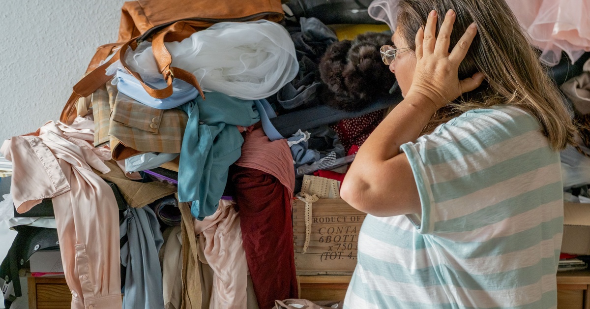 A woman holding her head as she stands in front of a huge pile of mixed clothes, jackets, bags, and fabrics in a home.