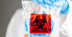A medical professional wearing protective gear and gloves holds up a plastic bag with a red label "Biohazard" on it.