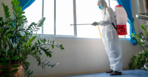 A person in a full-body protective suit with a spraying machine strapped to their back, sanitizing a home window area.