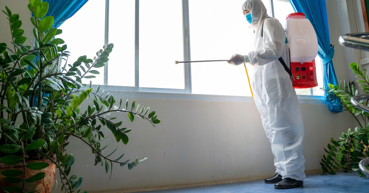 A person in a full-body protective suit with a spraying machine strapped to their back, sanitizing a home window area.