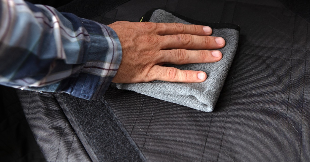 A hand wiping a car seat with a microfiber cloth, with seat stitching and interior trim visible nearby.