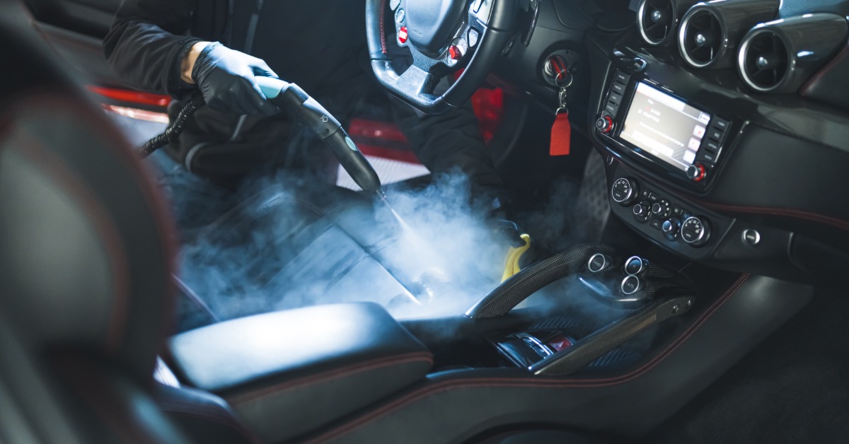 A worker using a steam cleaner on car seats inside a vehicle, with the dashboard and door panel visible.